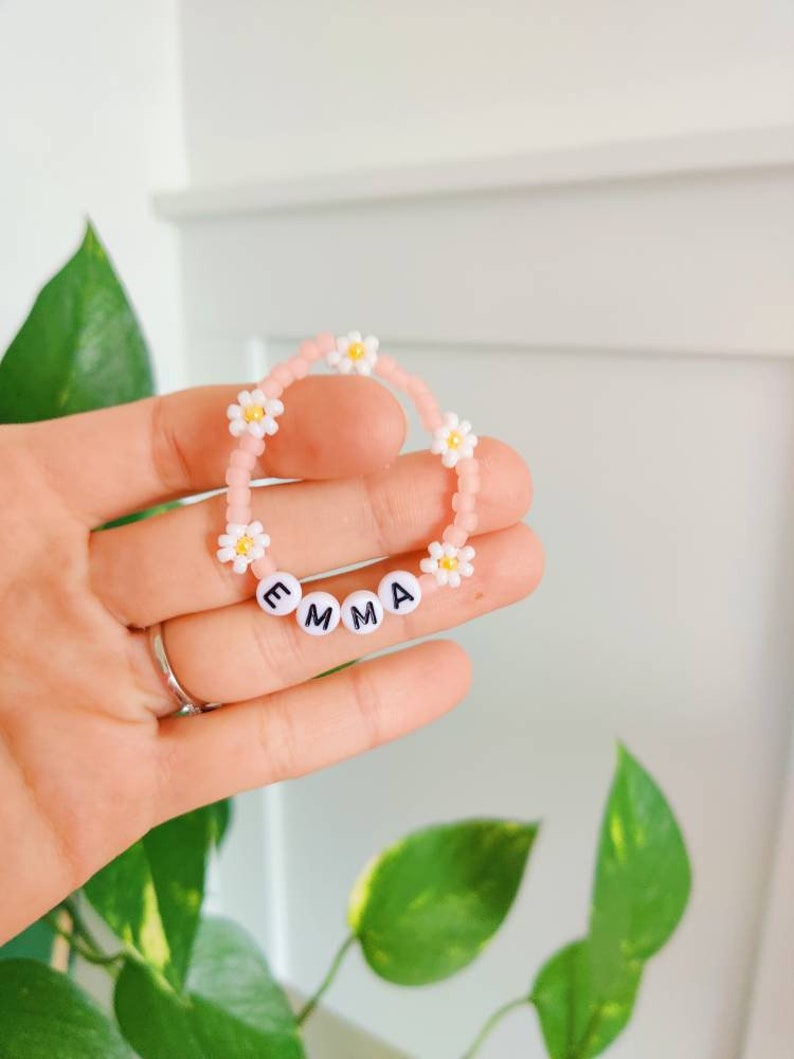 May include: Pink beaded bracelet with white daisy beads and white letter beads spelling the name "EMMA".