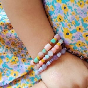 May include: A child's wrist adorned with four colourful beaded bracelets. The bracelets are made of various pastel-coloured beads, including pink, purple, turquoise, and yellow.