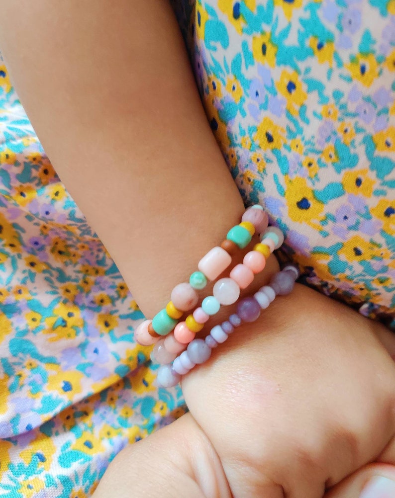 May include: A child's wrist adorned with four colourful beaded bracelets. The bracelets are made of various pastel-coloured beads, including pink, purple, turquoise, and yellow.