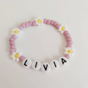 May include: A pink beaded bracelet with white daisy charms and white letter beads spelling out the name "LIVIA".