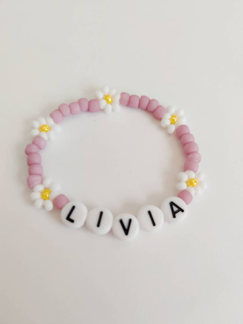 May include: A pink beaded bracelet with white daisy charms and white letter beads spelling out the name "LIVIA".