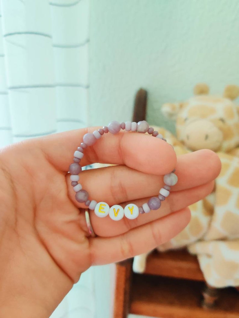 May include: A purple and white beaded bracelet with the name "EVY" spelled out in gold letters on white beads.
