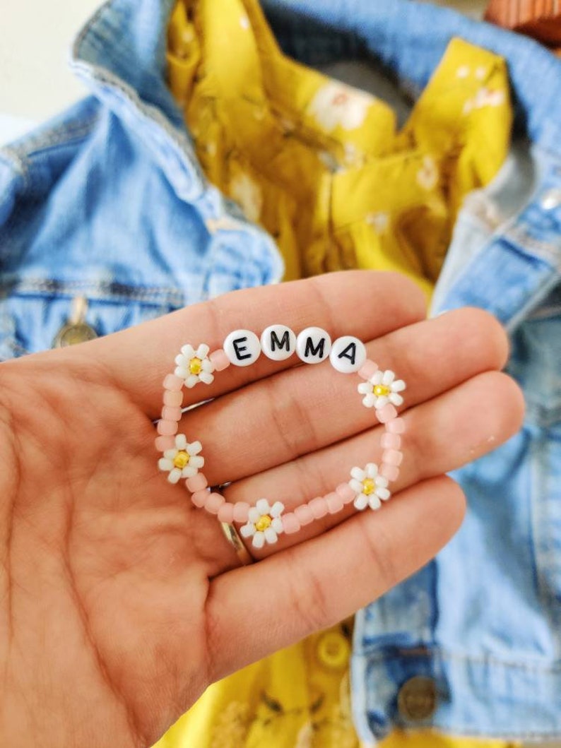May include: A pink beaded bracelet with white daisy beads spelling out the name "EMMA".