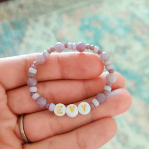 May include: A purple and white beaded bracelet with the name "EVY" spelled out in gold letters on white beads.
