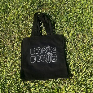 May include: Black tote bag with white text that reads "BASIC BRUJA" in a whimsical font. The bag has two handles and is sitting on a bed of green grass. A small flower and bat graphic are above the word "BRUJA".