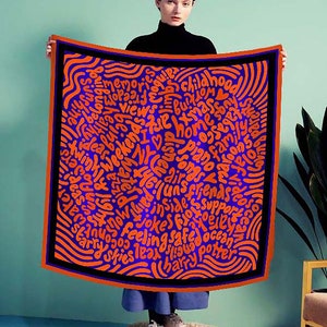May include: A woman is holding a blue and orange silk scarf with a black border. The scarf is covered in a pattern of orange words on a blue background. The words include "I love NY", "memories", "friends", "family", "ocean", "jokes", "safe", and "feeling".
