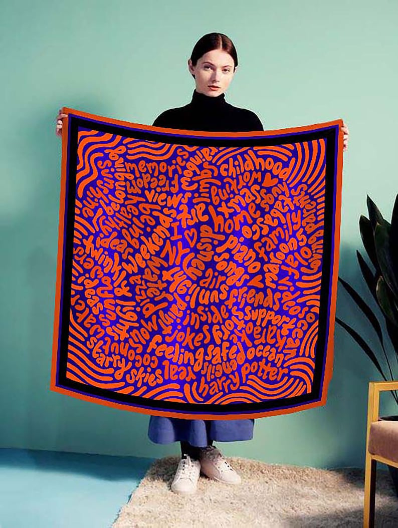 May include: A woman is holding a blue and orange silk scarf with a black border. The scarf is covered in a pattern of orange words on a blue background. The words include "I love NY", "memories", "friends", "family", "ocean", "jokes", "safe", and "feeling".