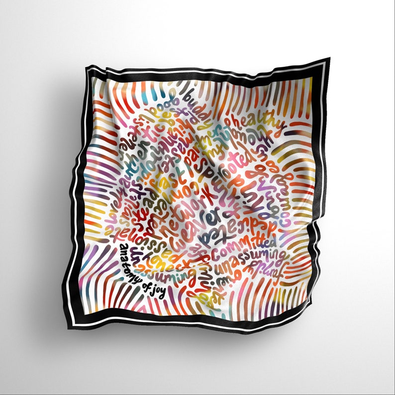 May include: A white silk scarf with a black border and a colorful design of words and swirls. The words include "authentic", "love", "joy", "community", "work", "healthy", "committed", "unassuming", and "blessed".