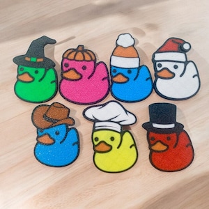 May include: Eight rubber duck figurines with different hats. The ducks are yellow, pink, green, blue, and red. The hats include a witch's hat, a cowboy hat, a chef's hat, a top hat, a Santa hat, and a beanie.
