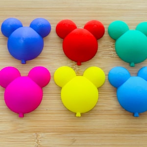 Op de afbeelding: Zes kleurrijke, ballonvormige decoraties in blauw, rood, groen, roze, geel en lichtblauw. Elk heeft drie kleinere cirkels bovenop, die op een muizenkop lijken. Ze zijn gerangschikt op een lichtbruin houten oppervlak.
