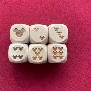 May include: Six wooden dice with etched Mickey Mouse head designs. Each die face features a different number of Mickey Mouse heads, ranging from one to six. The dice are arranged on a red surface.