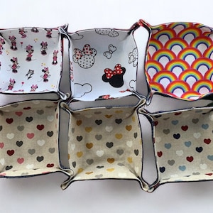 May include: Six fabric storage bins with different patterns. The bins are lined up in two rows of three. The patterns include hearts, rainbows, and a cartoon character.