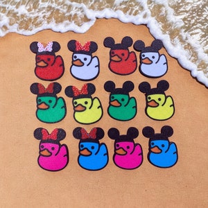 Może przedstawiać: Zestaw 12 gumowych kaczek z uszami Myszki Miki i w różnych kolorach, w tym czerwonym, białym, zielonym, żółtym, niebieskim i różowym. Kaczki są ułożone w siatkę na tle piaszczystej plaży.