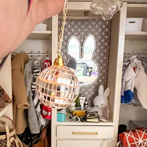 May include: A gold disco ball hanging from a chain with a white cord. The disco ball is made of small mirrored tiles and is reflecting light. The disco ball is hanging in a dollhouse closet with a mirror and other decorations.