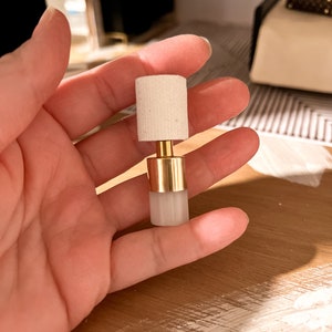 May include: A small, white and gold lamp with a round, white shade. The lamp is held in a hand.