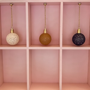 May include: Three hanging light fixtures with different colored shades. The shades are made of a woven material and are white, brown, and black. The light fixtures are hanging from a white shelf with three compartments.