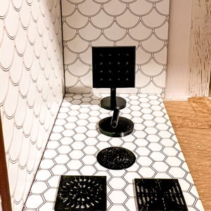 May include: Four black drain covers with different designs on a white hexagonal tile floor. The drain covers are square and round. The wall behind the drain covers is white with a pattern of overlapping half circles.