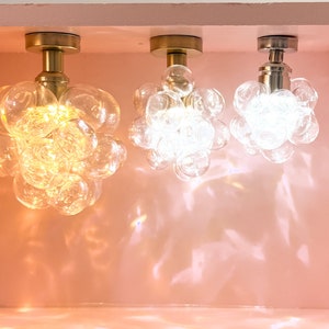 May include: Three modern ceiling lights with clear glass bubble shades and metallic fixtures. The lights are mounted on a pink surface, casting light and shadows. The fixtures have a warm, inviting glow.