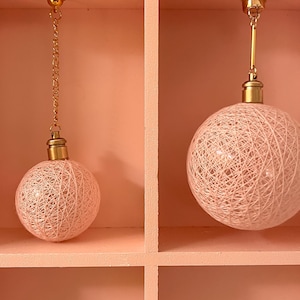May include: Two pink string lights hanging from a shelf. The lights are round and have a gold metal frame.