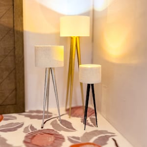 May include: Three tripod floor lamps with white fabric lampshades. The lamps are on a white floor with a patterned design. The lampshades are all different sizes and the lamps are made of different materials.