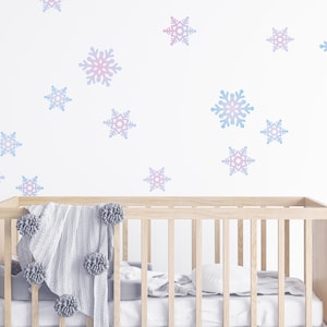May include: A wooden crib with a gray blanket and pom-pom accents. The white wall is decorated with blue and pink snowflake decals. The crib is filled with white bedding.