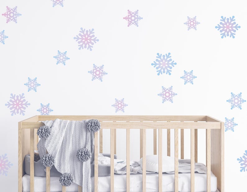 May include: A wooden crib with a gray blanket and pom-pom accents. The white wall is decorated with blue and pink snowflake decals. The crib is filled with white bedding.