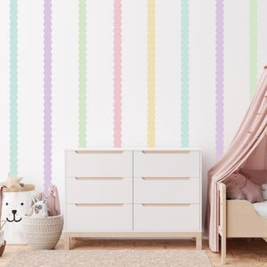 Può includere: Una cassettiera bianca con sei cassetti è posizionata davanti a un muro decorato con strisce verticali a festoni pastello gialle, blu, verdi, rosa e viola. Un lettino con baldacchino rosa è sulla destra.