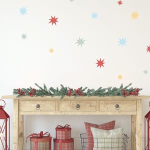 May include: A festive scene with a wooden console table adorned with a garland of greenery and red berries. Red lanterns flank the table, and gifts are stacked below. Colorful star decals decorate the white wall, creating a cheerful holiday ambiance.