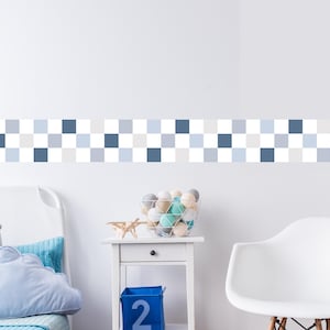Op de afbeelding: Een witte muur met een blauwe, grijze en witte geblokte rand. De kamer heeft een wit bed met blauwe kussens, een wit nachtkastje met een mand met garenballen en een blauwe opbergdoos met het nummer 2.