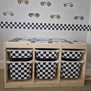 May include: A wooden toy storage unit with four black and white checkered storage bins. The unit has a top surface with a printed race track design. The wall behind the unit has a black and white checkered border and several black, gray, and brown toy car decals.