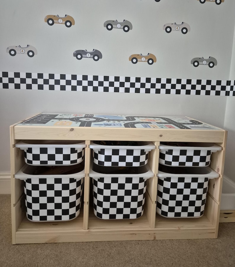 May include: A wooden toy storage unit with four black and white checkered storage bins. The unit has a top surface with a printed race track design. The wall behind the unit has a black and white checkered border and several black, gray, and brown toy car decals.