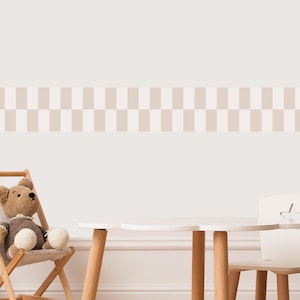 Op de afbeelding: Een kinderkamer met een witte tafel en stoelset met houten poten. Een teddybeer zit in een houten klapstoel. Een decoratieve rand met een geblokt patroon in beige en gebroken wit siert de muur.