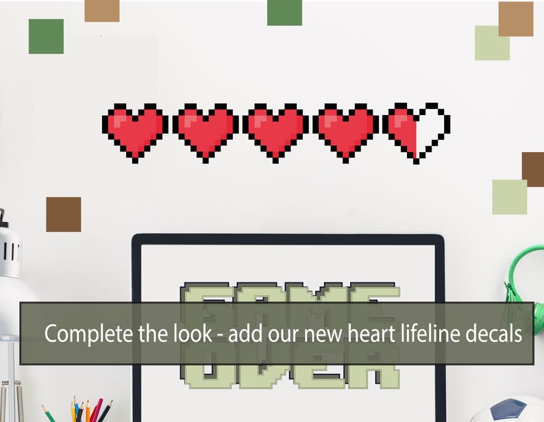 Pu&ograve; includere: Un muro bianco con una fila di cuori pixelati rossi, un monitor di computer con la scritta "GAME OVER" sullo schermo e il testo "Completa il look - aggiungi i nostri nuovi adesivi di linea di vita a forma di cuore".