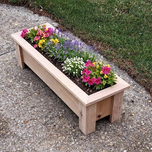 May include: A rectangular wooden planter box filled with colorful flowers. The flowers include pink, yellow, purple, and white blooms. The planter is made of light-colored wood and sits on a concrete surface next to green grass.