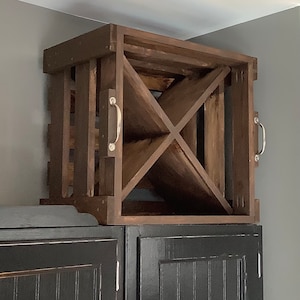 May include: A dark brown wooden wine rack with a criss-cross design. The rack is mounted on top of two black cabinets.