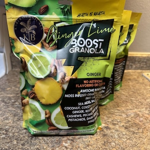 Puede incluir: Tres bolsas de granola Ginger Lime Boost. Las bolsas son verdes y amarillas con imágenes de limas, jengibre y nueces. El texto en las bolsas incluye "Ginger Lime Boost Granola" y "Sin saborizantes ni colorantes artificiales."
