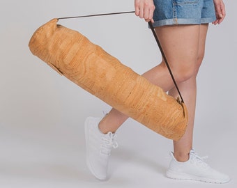 May include: A woman wearing a white t-shirt and denim overalls carries a cork yoga mat with a brown strap.