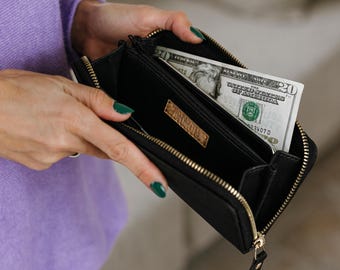 May include: A black zippered wallet with a gold-toned metal zipper and a rectangular gold-toned brand tag inside. The wallet is open and contains a twenty-dollar bill from the United States, clearly showing the text Twenty Dollars.