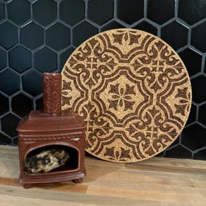 May include: A brown ceramic miniature fireplace with a decorative cork trivet. The trivet has a brown, ornate pattern. The fireplace has a small opening with a decorative object inside. The background features black hexagonal tiles.