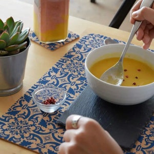May include: A white ceramic bowl filled with yellow soup topped with red peppercorns, placed on a rectangular black slate coaster. The bowl sits on a blue and white patterned placemat alongside a small glass dish of peppercorns.