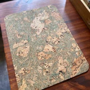 May include: Rectangular cork placemat with rounded corners. The surface features a natural, mottled pattern of beige, brown, and green hues. The placemat is designed for use on a table or desk.