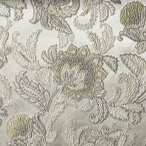 Puede incluir: Primer plano de una tela blanca con un patrón floral plateado y dorado. La tela tiene un aspecto texturizado y tejido.