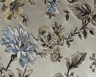 May include: Close-up of a fabric with an embroidered floral pattern. The design features large, detailed flowers in shades of blue, gold, and brown, set against a light, neutral background. The embroidery creates a textured, three-dimensional effect.