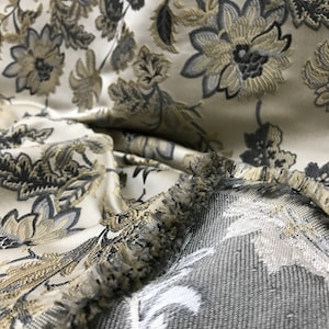 May include: Close-up of a white fabric with a floral pattern in shades of gray and gold. The fabric has a fringe border.