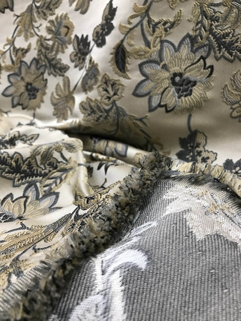 May include: Close-up of a white fabric with a floral pattern in shades of gray and gold. The fabric has a fringe border.