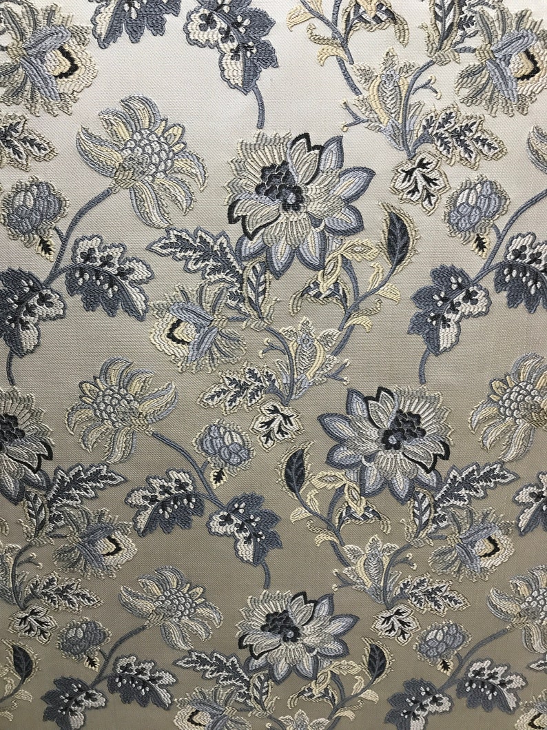 May include: A beige fabric with a repeating pattern of blue and white floral designs. The fabric has a subtle sheen and a textured surface.
