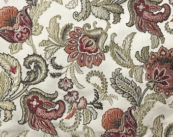 May include: Close-up of a cream-colored fabric with an intricate floral pattern. The design features large, stylized flowers in shades of burgundy, rust, and gold, with green and olive-colored leaves and stems. The fabric has a textured appearance.