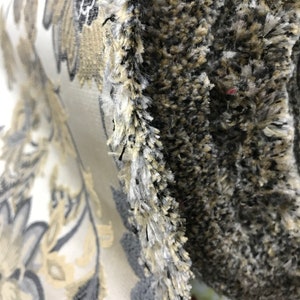 May include: Close-up of a fabric with a floral pattern in shades of beige, gray, and white. The fabric has a textured surface and a fringe edge.