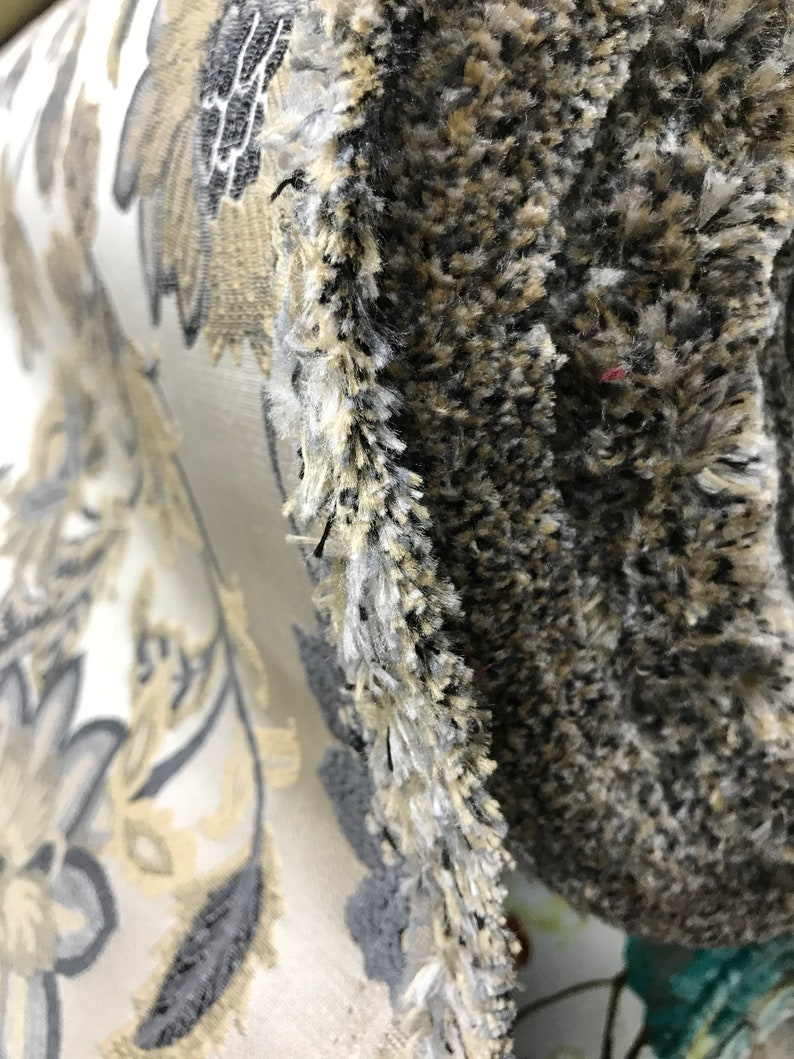 May include: Close-up of a fabric with a floral pattern in shades of beige, gray, and white. The fabric has a textured surface and a fringe edge.