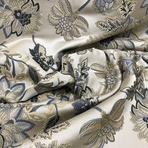 May include: Close-up of a white fabric with a floral pattern. The fabric has a raised texture and features gray and beige flowers.